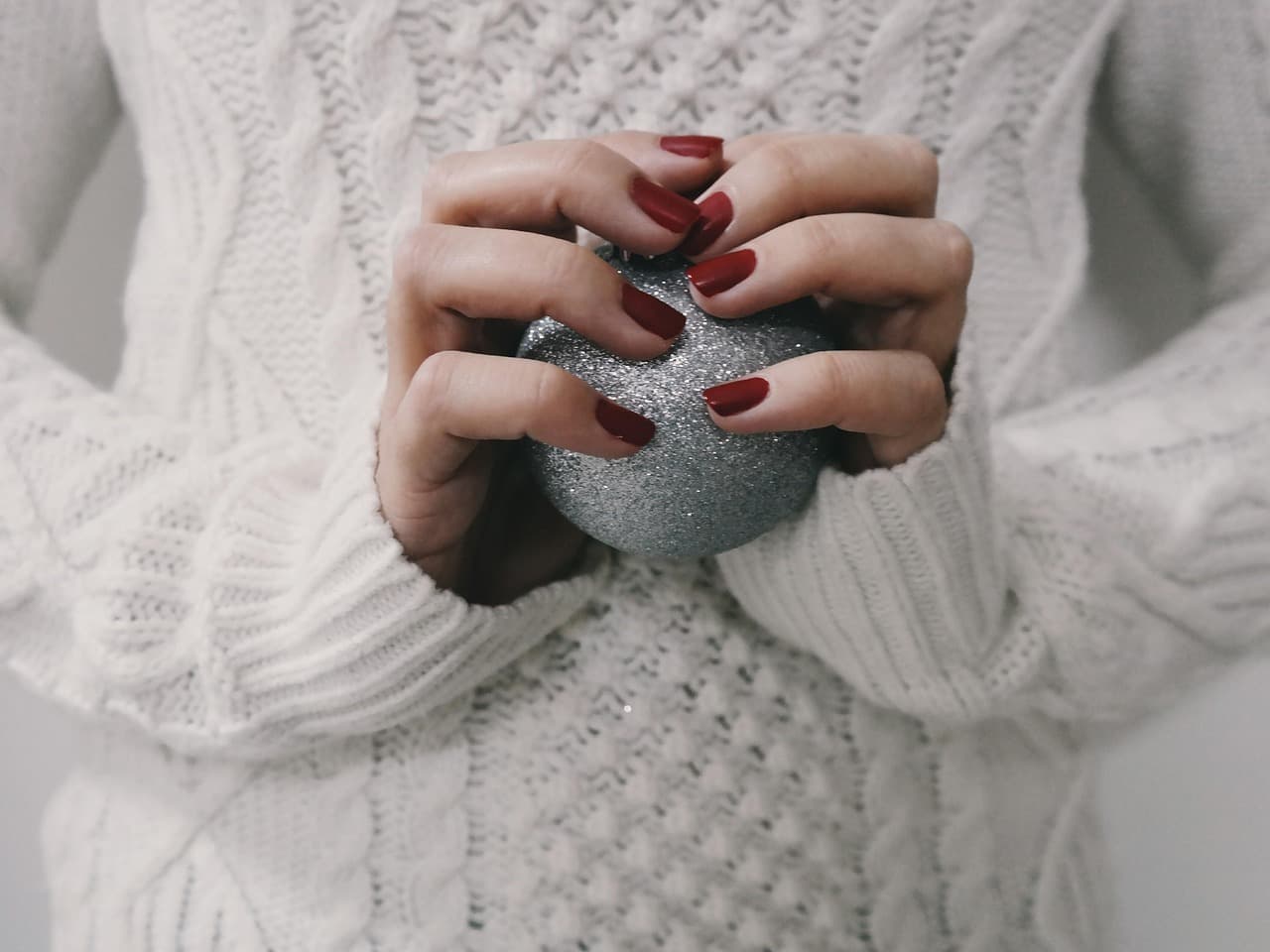 Deep red nails on hands in a cream knit, holding a silver bauble